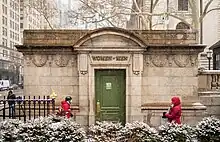 A stone building with decorative carvings near the roof line.  A single green door is labeled "WOMEN – MEN" above the door.