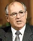 The image is a portrait of a middle-aged man (Mikhail Gorbachev) with short, light-colored hair, wearing large-framed glasses and a dark suit with a light-colored shirt and a dark tie. He has a noticeable red birthmark on his forehead. The background of the photo is indistinct.