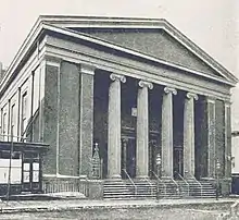 Black and white photograph of a building with columns, stairs leading to the street, and a neoclassical pediment