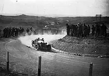 An old sports car drives on unsealed roads as people stand on hills around the road and watch