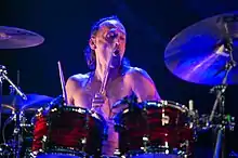 Sebastian Thomson drumming shirtless against a black background with blue stage lighting