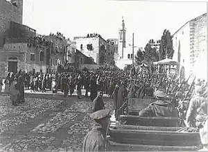 Towers and buildings some with people looking down from roofs with local people and soldiers in a large square with motor car in foreground