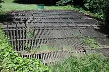 A horizontal wooden rack roughly 30 feet (9.1 m) wide and 30 feet (9.1 m) long lies in flat, grassy area.