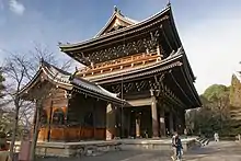 A large two-storied wooden gate with a veranda on the upper story and a hip-and-gable style roof. A small wooden building with gabled roof is placed next to it.