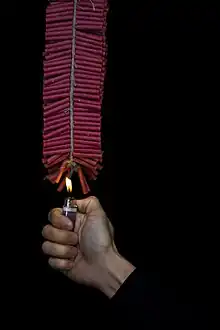 A string of red firecrackers. A young man holds a flame to set it off.