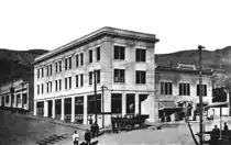A black and white photograph of the recently opened Cook Bank building in 1908 from the diagonally opposite corner of the intersection. The bank is a three-story white masonry building with many windows on the two visible sides and a door on each side. A cornice wraps around the building at the roofline; the roof is flat. About twelve people are in the street outside the building, along with a car, and three telephone utility poles. A two-story building with awnings is directly attached to the longer side of the bank building (to the right). The road slopes upward to the left, passing a single story building. Hills are visible in the background.