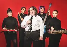 Fatty Cakes and the Puff Pastries standing in front of a red back drop. Crow, Audrey, Vishinna, and Stacey are wearing all Black with silver accents with a playful but serious look. Amber is standing in the center with a white dress shirt and black pants and a black tie, holding a microphone and smiling.