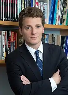 Nisbet standing in a business suit with arms crossed in front of bookshelves