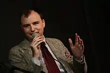 Photo of Jesse Thorn speaking into a microphone. He is caucasian and wearing a coat and tie.