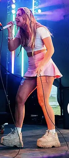 Side-on view of a blonde woman in a white crop top and pink miniskirt wearing a crocheted frog hat, white socks, and white chunky trainers, holding a microphone to her mouth with her right hand and its cord with her left.