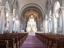 Looking down central aisle with pews on either side; line of pillars connected at tops by arches on either side; altar under half-dome at far end