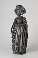 Figure F: Woman with crossed hands and a shaved hairline. She is dressed in a turban decorated with vertical rows of pearls, and a large jewel at front.