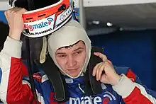 A man in his early twenties wearing a white fireproof balaclava around most of his head and dark red, blue and white racing overalls. He is putting on his helmet and HANS device.