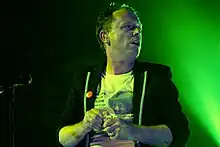 Torquil Campbell wearing a white print t-shirt and a black hoodie, backlit in green in front of a microphone onstage, looking left of camera