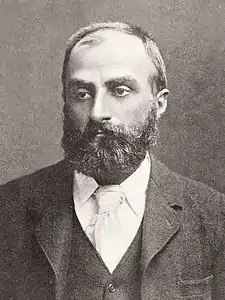 Heavily bearded man with dark receding hair, wearing a dark coloured jacket, white-collar and pale tie. He is looking slightly to the left, with a solemn expression