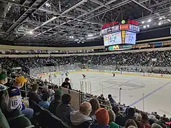 A 3/4 view of an ice hockey rink. The scoreboard says HEB Center and has a close-up shot of a hockey goalie. There are two teams on the ice, one in green and white and the other in maroon.