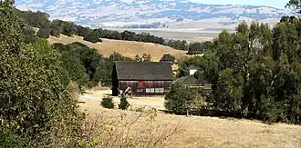 A weathered old barn sits among hills, trees, and dry grass